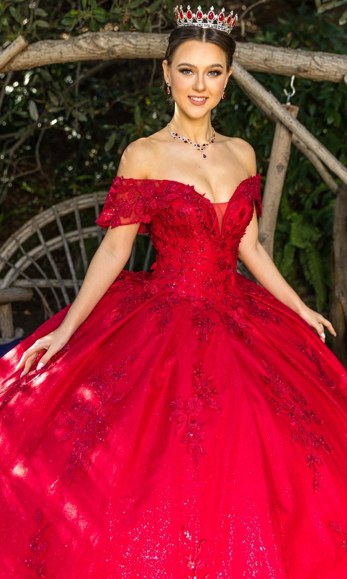 A woman wearing the Quinceanera Ball Gown 8045J with sparkling red glitter lace embroidery and a tiara stands outdoors before a wooden arch and greenery.